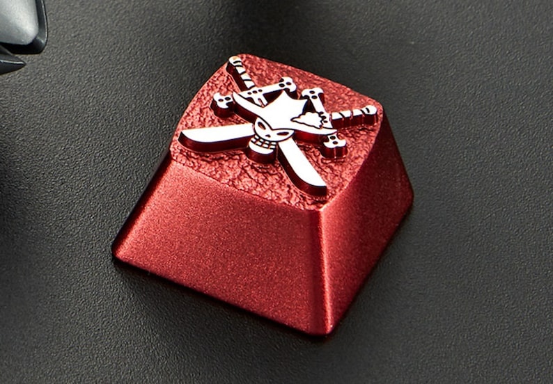 May include: A red metal keycap with a silver skull and crossbones design. The skull is wearing a hat and the crossbones are crossed swords.