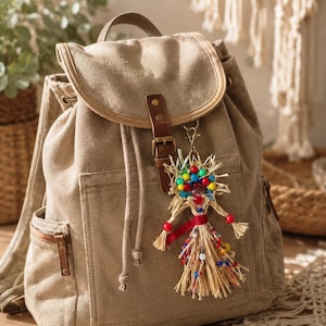 Op de afbeelding: Een beige canvas rugzak met een bruine leren riemsluiting en een decoratieve strooien poppen-sleutelhanger met kleurrijke kralen, een rood lint en een metalen karabijnhaak die aan de voorflap van de tas hangt.