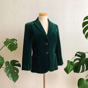 Peut inclure: Un blazer en velours vert foncé avec un revers cranté et une fermeture à deux boutons. Le blazer a deux poches avant et est présenté sur un mannequin. La veste est de couleur unie et a des manches longues.