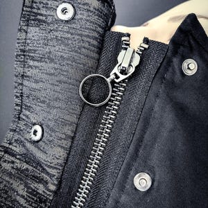 May include: Close-up of a black jacket with a silver zip and a silver ring pull. The jacket has a textured fabric and a silver button.