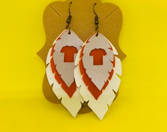 May include: A pair of dangle earrings with a white and orange feather design. The earrings feature a small t-shirt charm.