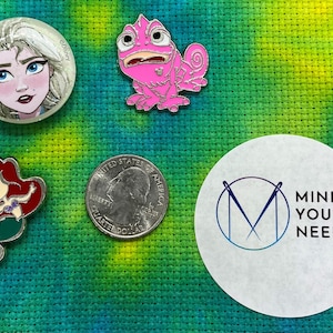 Puede incluir: Una colección de cinco pines de esmalte con personajes de Disney. Los pines están sobre un fondo de tela verde y amarillo. Los pines incluyen un camaleón rosa, una sirena con cabello rojo, una princesa con cabello rubio y una moneda de 25 centavos.