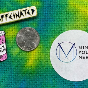 Könnte beinhalten: Drei Emaille-Pins auf einem grün-gelben Stoffhintergrund. Die Pins sind ein weiß-goldener Pin mit der Aufschrift "caffeinated", eine rosa-weiße Kaffeetasse mit dem Text "Mermaid's Macchiato" und ein weißer Kreis mit dem Text "Mind Your Needles" und einem stilisierten Nadel- und Faden-Design.