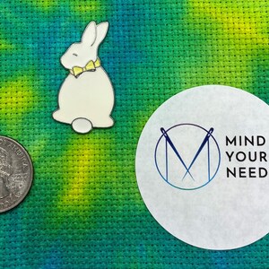 May include: White ceramic bunny with a yellow bow tie, sitting on a green and blue fabric background. The bunny is next to a white circle with the text "MIND YOUR NEEDLES" in black.