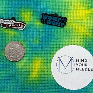 May include: Three enamel pins with humorous phrases: "Sounds Like BULLSHIT to Me!" and "WOMP WOMP" in black and blue. A US quarter coin and a white sticker with the text "MIND YOUR NEEDLES" are also visible.