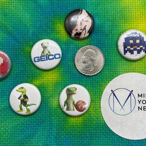 May include: A collection of eight round buttons with various designs. The buttons feature images of a gecko, a ghost, a video game character, and a woman's face. One button has the text "GEICO" on it. Another button has the text "MIND YOUR NEEDLES" on it. The buttons are arranged on a green and yellow tie-dye fabric.