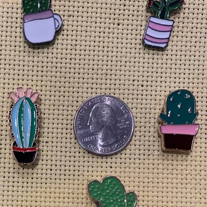 May include: Five enamel cactus pins in different styles. The pins are all gold-toned and feature different colored pots and cacti. The pins are arranged in a grid pattern on a beige fabric background.