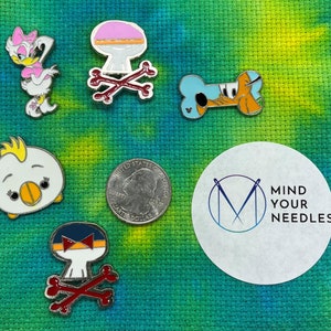 May include: A collection of enamel pins featuring various cartoon characters, including a duck, a dog, and a mushroom. The pins are displayed on a green and yellow fabric background with a white sticker that says "Mind Your Needles".