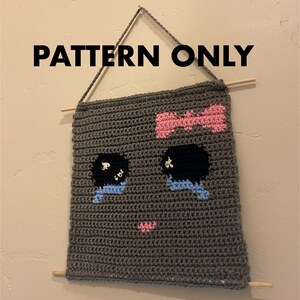 May include: A gray crochet wall hanging with a cartoon face featuring black eyes, blue tears, and a pink bow. The text "PATTERN ONLY" is printed above the hanging.
