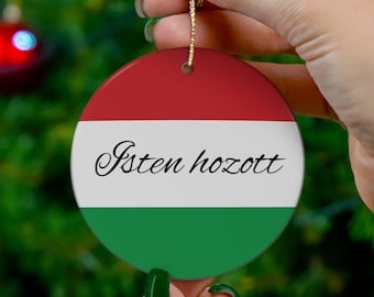Puede incluir: Un adorno navideño redondo con el diseño de la bandera húngara, con rayas rojas, blancas y verdes. El adorno tiene el texto "Isten hozott". Se sostiene frente a un árbol de Navidad con adornos rojos.