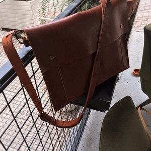 May include: A brown leather messenger bag with a long adjustable strap. The bag has a flap closure with a metal snap and a front pocket. The bag is hanging on a black metal railing.