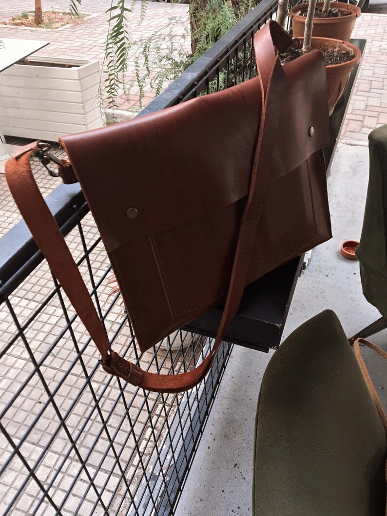 May include: A brown leather messenger bag with a long adjustable strap. The bag has a flap closure with a metal snap and a front pocket. The bag is hanging on a black metal railing.