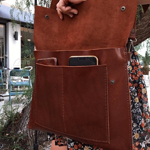 May include: A brown leather messenger bag with a flap closure and a shoulder strap. The bag has a large front pocket and a smaller pocket inside. A smartphone is visible inside the bag. The bag is open, revealing the interior.