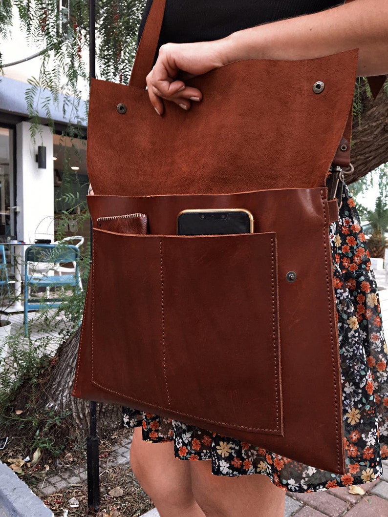 May include: A brown leather messenger bag with a flap closure and a shoulder strap. The bag has a large front pocket and a smaller pocket inside. A smartphone is visible inside the bag. The bag is open, revealing the interior.