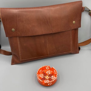 May include: A brown leather messenger bag with a flap closure and a shoulder strap. A small, round, orange ashtray with a white and red floral design is in the foreground.