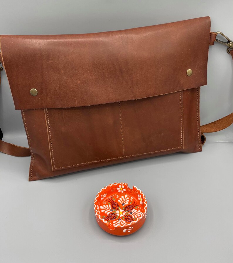 May include: A brown leather messenger bag with a flap closure and a shoulder strap. A small, round, orange ashtray with a white and red floral design is in the foreground.
