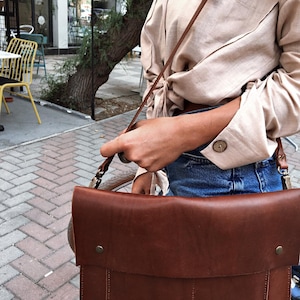 May include: A brown leather messenger bag with a flap closure and a shoulder strap. The bag has a rectangular shape with visible stitching and metal hardware. The bag is carried over the shoulder.