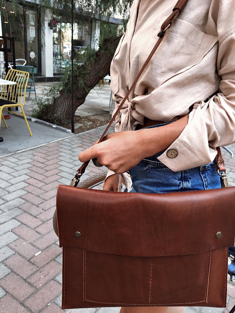May include: A brown leather messenger bag with a flap closure and a shoulder strap. The bag has a rectangular shape with visible stitching and metal hardware. The bag is carried over the shoulder.