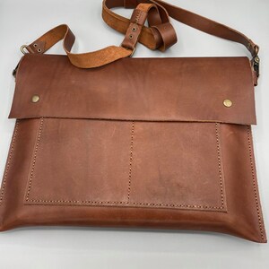 May include: A brown leather messenger bag with a flap closure and adjustable shoulder strap. The bag features a front pocket and is secured with a brass-colored snap. The leather has visible grain and stitching details.