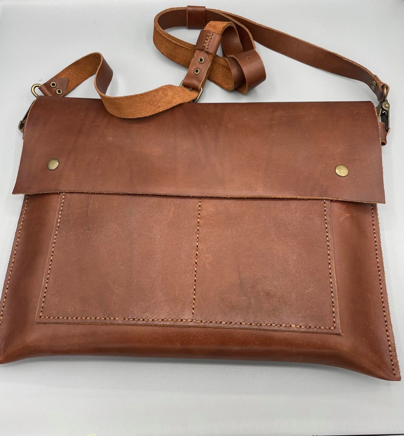 May include: A brown leather messenger bag with a flap closure and adjustable shoulder strap. The bag features a front pocket and is secured with a brass-colored snap. The leather has visible grain and stitching details.