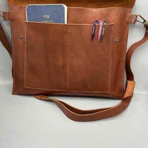 May include: A brown leather messenger bag with a flap closure, shoulder strap, and a front pocket. Inside the bag, a small blue notebook and three pens are visible. The bag is made of a rich, textured leather.