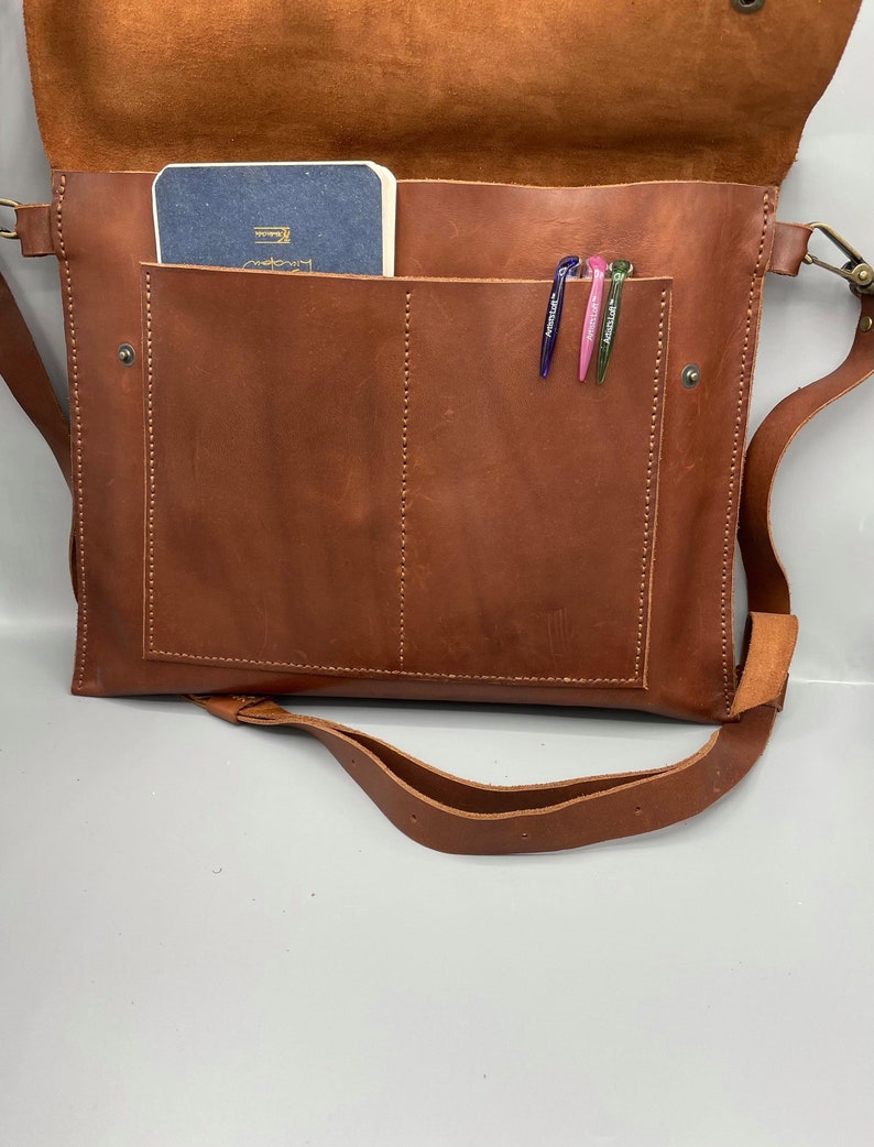 May include: A brown leather messenger bag with a flap closure, shoulder strap, and a front pocket. Inside the bag, a small blue notebook and three pens are visible. The bag is made of a rich, textured leather.