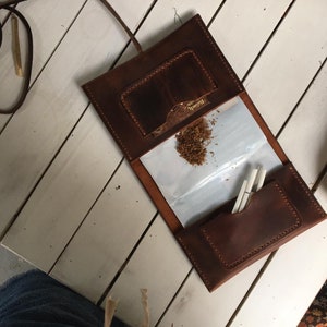 Puede incluir: Una bandeja de enrollado de cuero marrón con una solapa que se abre para revelar un papel blanco y una pequeña bolsa. La bandeja tiene un pequeño bolsillo en el exterior con la palabra "Brownie" impresa en él.
