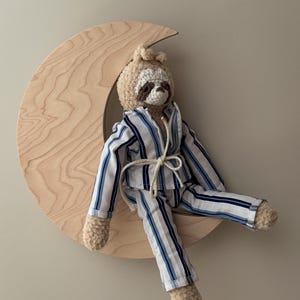 Könnte beinhalten: Ein Plüschfaultier in gestreiftem Pyjama, das auf einem hölzernen Halbmond sitzt. Der Pyjama ist weiß mit blauen Streifen. Das Faultier hat eine gehäkelte Kopfbedeckung und besteht aus weichem, strukturiertem Material.