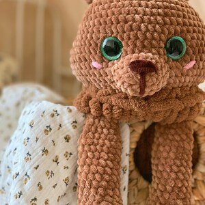Puede incluir: Un oso de peluche de crochet marr&oacute;n con ojos verdes y nariz rosa. El oso est&aacute; sentado en una cesta tejida y tiene los brazos extendidos.
