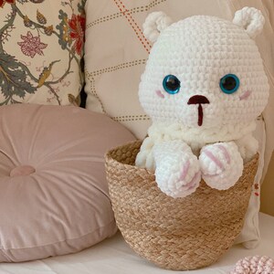 Puede incluir: Un oso de peluche blanco de crochet con ojos azules se sienta en una cesta tejida. La cesta est&aacute; en una cama blanca con una manta rosa y una almohada rosa.