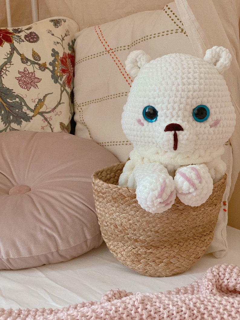 Puede incluir: Un oso de peluche blanco de crochet con ojos azules se sienta en una cesta tejida. La cesta est&aacute; en una cama blanca con una manta rosa y una almohada rosa.