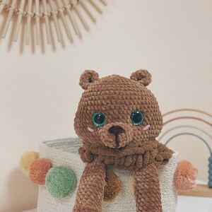 Puede incluir: Un oso de peluche de crochet marr&oacute;n con ojos verdes se sienta en una cesta tejida blanca con pompones de colores.