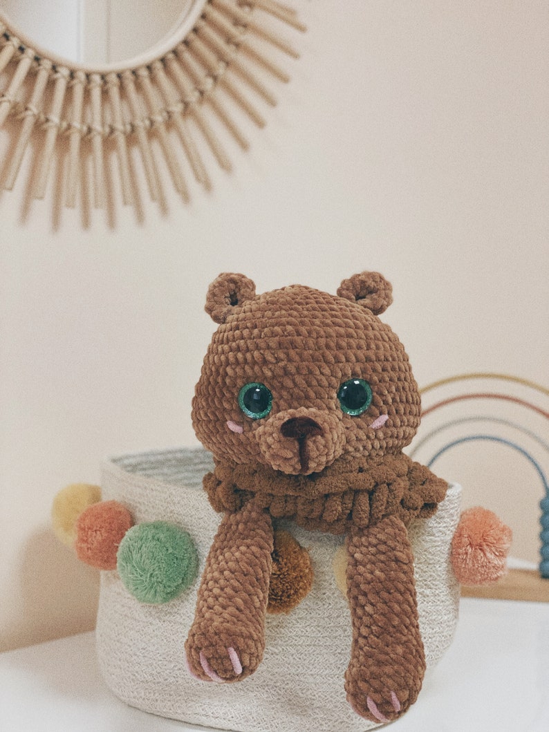Puede incluir: Un oso de peluche de crochet marr&oacute;n con ojos verdes se sienta en una cesta tejida blanca con pompones de colores.