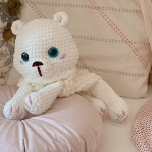 Puede incluir: Oso de peluche blanco tejido a crochet con ojos azules y nariz rosa. El oso est&aacute; sentado en una almohada rosa.