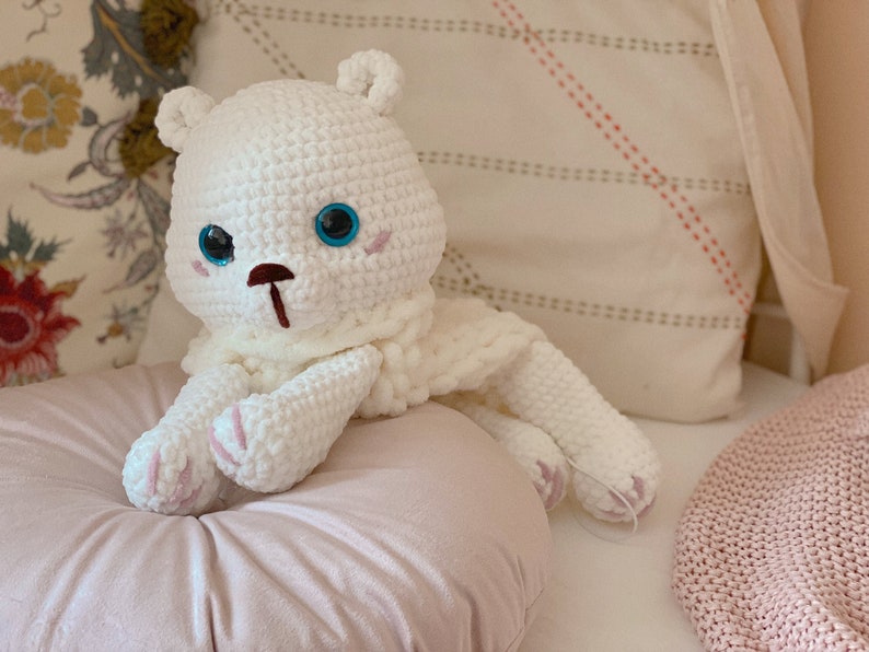 Puede incluir: Oso de peluche blanco tejido a crochet con ojos azules y nariz rosa. El oso est&aacute; sentado en una almohada rosa.
