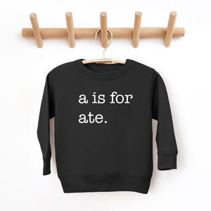 May include: Black sweatshirt with the text "a is for ate" in white. The sweatshirt is hanging on a wooden coat rack against a white background. A casual, comfortable garment.
