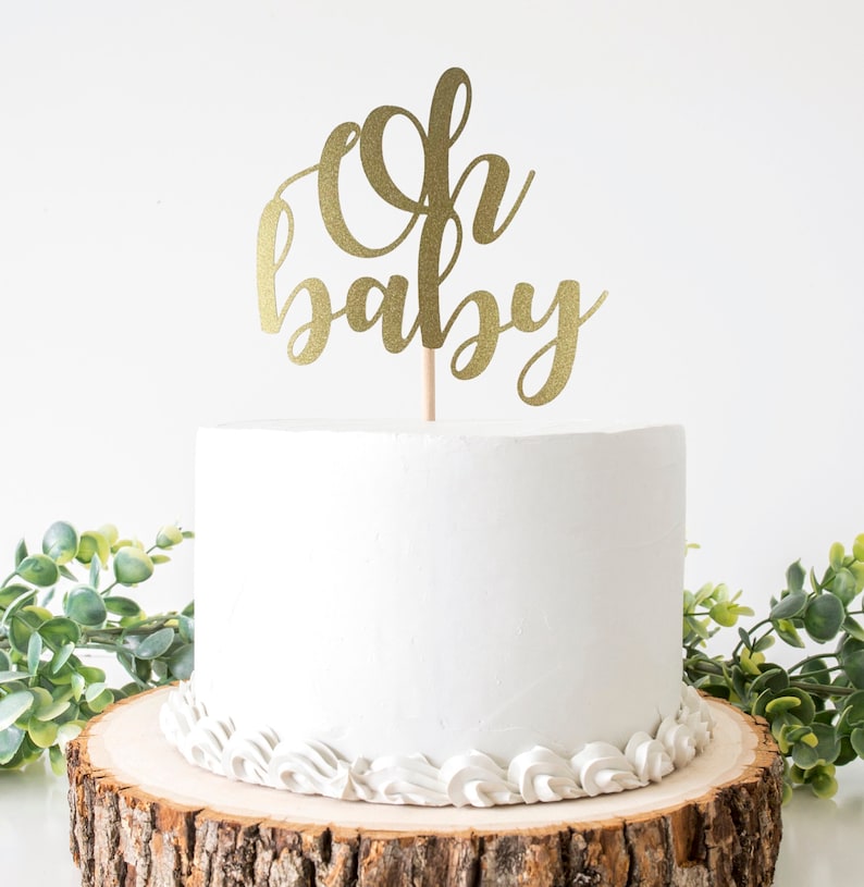 May include: A white frosted cake with a gold glitter "Oh baby" cake topper. The cake sits on a wooden cake stand with a decorative white frosting border. Greenery is placed around the cake.