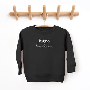 May include: Black crewneck sweatshirt with the words "kuya hendrix" in white script. The sweatshirt is hanging on a wooden hanger against a white background. The sweatshirt is a children's apparel item.