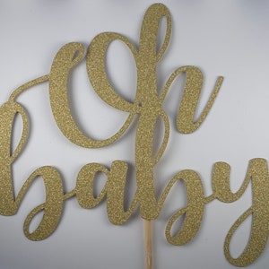 May include: A gold glitter "Oh Baby" cake topper. The script lettering is attached to a wooden stick for easy insertion into a cake. The topper is designed for a baby shower or gender reveal party.