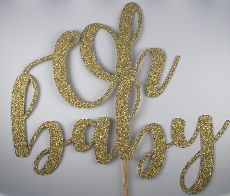 May include: A gold glitter "Oh Baby" cake topper. The script lettering is attached to a wooden stick for easy insertion into a cake. The topper is designed for a baby shower or gender reveal party.