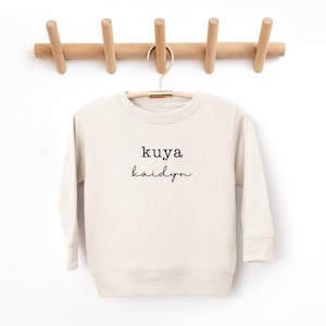 May include: A cream colored sweatshirt with the text "kuya kaidyn" printed in black.