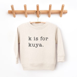 May include: A beige long-sleeved sweatshirt with the text "k is for kuya." printed in black.