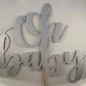 May include: Silver glitter "Oh Baby" cake topper on a wooden stick. The script lettering is in a cursive font. The cake topper is a decorative element for a baby shower or gender reveal party.