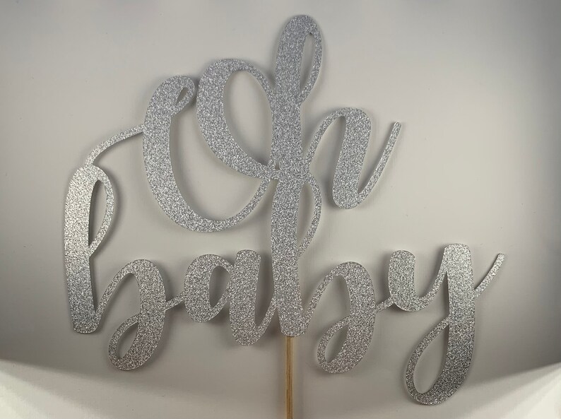 May include: Silver glitter "Oh Baby" cake topper on a wooden stick. The script lettering is in a cursive font. The cake topper is a decorative element for a baby shower or gender reveal party.