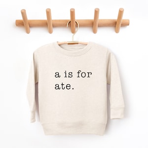 May include: A beige long-sleeved sweatshirt with the text "a is for ate." printed in black.