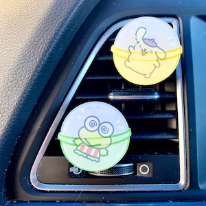 May include: Two round car air freshener clips with cartoon characters. One features a yellow bunny wearing a beret and the other features a green frog with a red and white striped shirt.