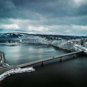 May include: An aerial view of a bridge over a lake in a snowy landscape. The bridge is gray and has a car driving across it. The lake is dark blue and the surrounding hills are covered in snow.