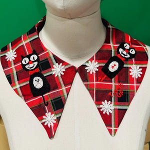 May include: A decorative collar with a red plaid pattern, featuring embroidered black cat and snowflake designs. The collar has a pointed shape and is displayed on a mannequin. The cat has a red bow tie.