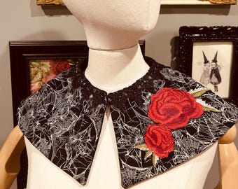 Könnte beinhalten: Ein schwarz-weißes Kragenmuster mit Spinnennetz-Muster und schwarzem Spitzensaum. Zwei rote Rosenapplikationen sind auf den Kragen genäht. Der Kragen wird auf einer Schaufensterpuppe präsentiert.
