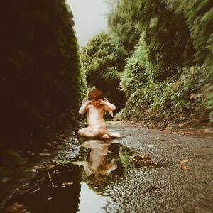 Puede incluir: Una estatua de una figura alada sentada en un charco de agua en un camino en un bosque. La figura está llorando y se cubre la cara con las manos.
